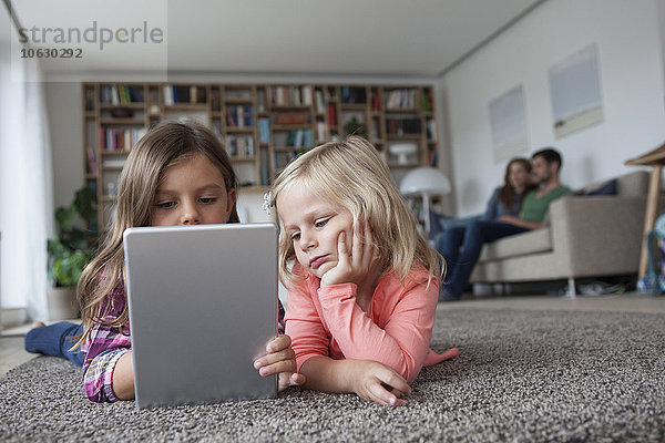Zwei kleine Schwestern liegen auf dem Boden im Wohnzimmer mit digitalem Tablett  während ihre Eltern im Hintergrund sitzen.