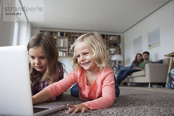 Zwei kleine Schwestern liegen auf dem Boden im Wohnzimmer mit Laptop  während ihre Eltern im Hintergrund sitzen.