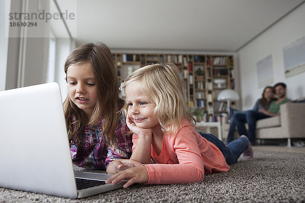 Zwei kleine Schwestern liegen auf dem Boden im Wohnzimmer mit Laptop  während ihre Eltern im Hintergrund sitzen.