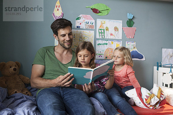 Vater und seine kleinen Töchter sitzen zusammen auf dem Bett im Kinderzimmer und lesen ein Buch.
