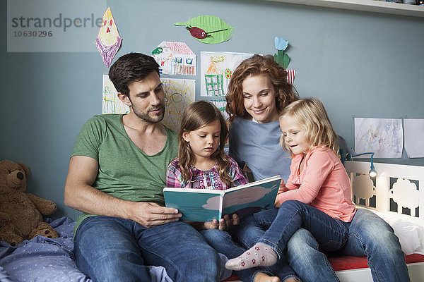 Paar und kleine Töchter sitzen zusammen auf dem Bett im Kinderzimmer und lesen ein Buch.