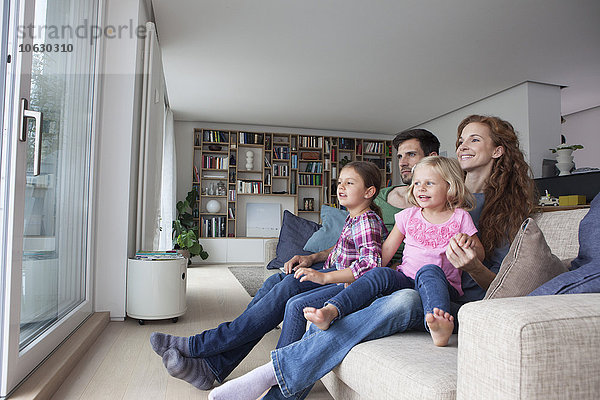 Paar und zwei kleine Mädchen auf der Couch im Wohnzimmer mit Blick durch die Terrassentür