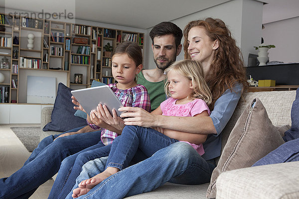 Paar sitzend mit ihren zwei kleinen Töchtern auf der Couch im Wohnzimmer mit Blick auf ein digitales Tablett.