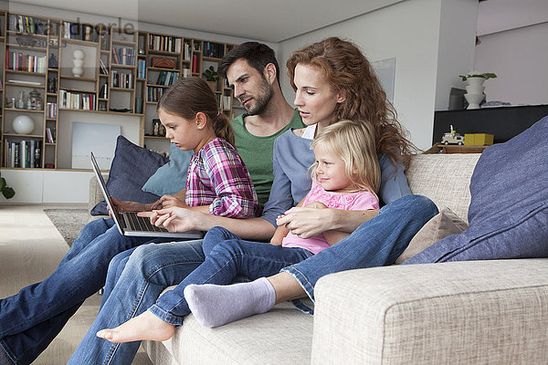 Paar sitzend mit ihren zwei kleinen Töchtern auf der Couch im Wohnzimmer mit Blick auf Laptop