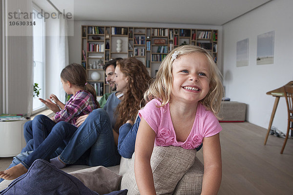 Porträt eines lächelnden kleinen Mädchens auf der Rückenlehne der Couch mit ihrer Familie im Hintergrund.