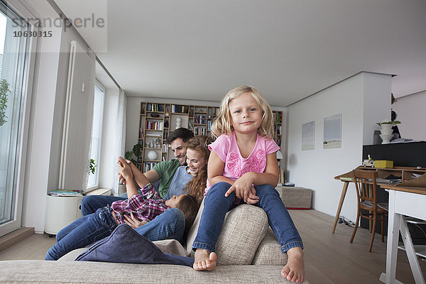 Porträt eines kleinen Mädchens  das auf der Rückenlehne der Couch sitzt  mit seiner Familie im Hintergrund.