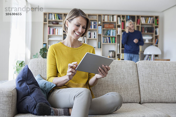 Lächelnde Frau sitzt auf der Couch mit digitalem Tablett  während ihr Mann im Hintergrund telefoniert.