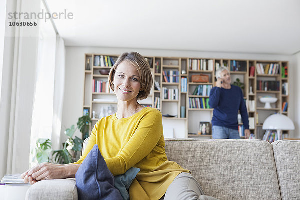 Lächelnde Frau sitzt auf der Couch  während ihr Mann im Hintergrund telefoniert.
