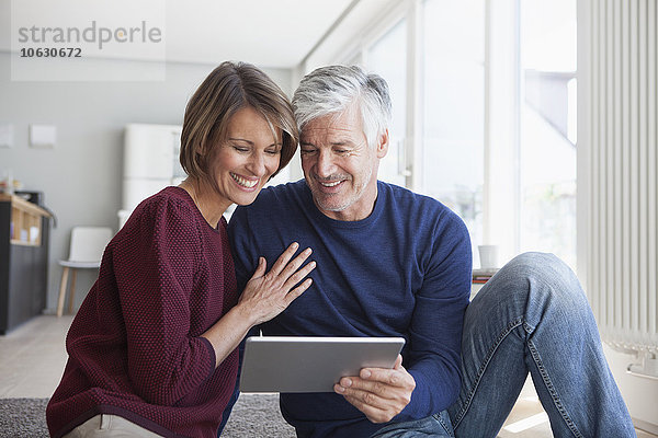 Lächelndes Paar sitzt zu Hause auf dem Boden und schaut auf ein digitales Tablett.