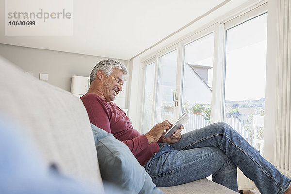 Lächelnder Mann sitzt zu Hause auf der Couch und benutzt ein digitales Tablett.