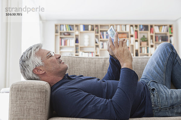 Erwachsener Mann  der zu Hause auf der Couch liegt und ein digitales Tablett benutzt.