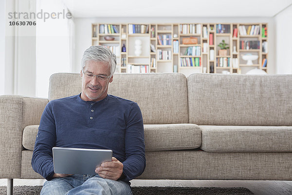 Lächelnder Mann sitzt zu Hause vor der Couch und benutzt ein digitales Tablett.