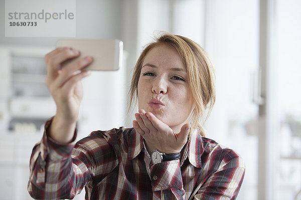 Junge Frau bläst einen Kuss  während sie einen Selfie mit ihrem Smartphone nimmt.