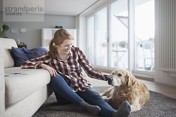 Lächelnde junge Frau sitzt neben ihrem Hund auf dem Boden zu Hause.