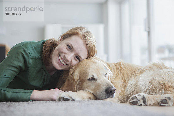 Porträt einer lächelnden jungen Frau  die mit ihrem Hund auf dem Teppich liegt.