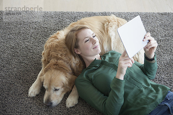 Lächelnde junge Frau  die mit ihrem Hund auf dem Teppich liegt.