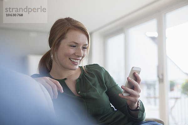 Porträt einer lächelnden jungen Frau auf der Couch mit Blick auf das Smartphone