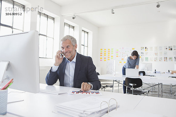 Lächelnder Geschäftsmann im Büro am Schreibtisch auf dem Handy