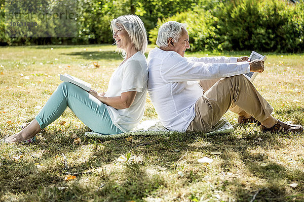 Ältere Paare sitzen Rücken an Rücken im Gras und lesen Bücher.