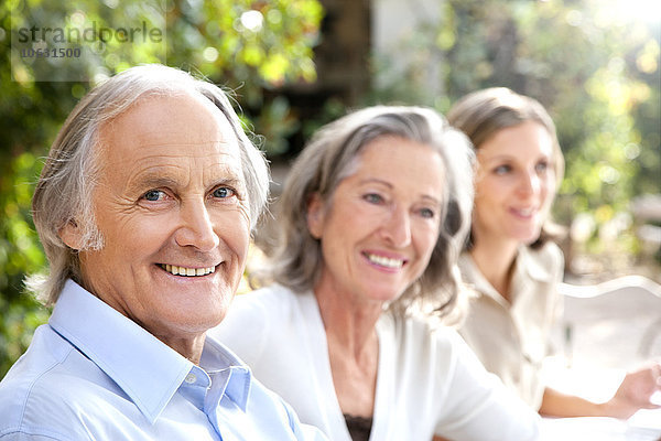 Porträt eines lächelnden älteren Mannes und zweier Frauen im Hintergrund  die im Garten sitzen.