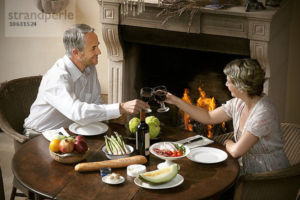Ein reifes Paar sitzt am gedeckten Tisch und toastet mit Rotwein vor offenem Feuer.