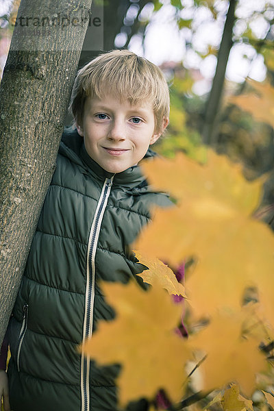 Porträt eines lächelnden Jungen  der sich im Herbst an den Baumstamm lehnt.