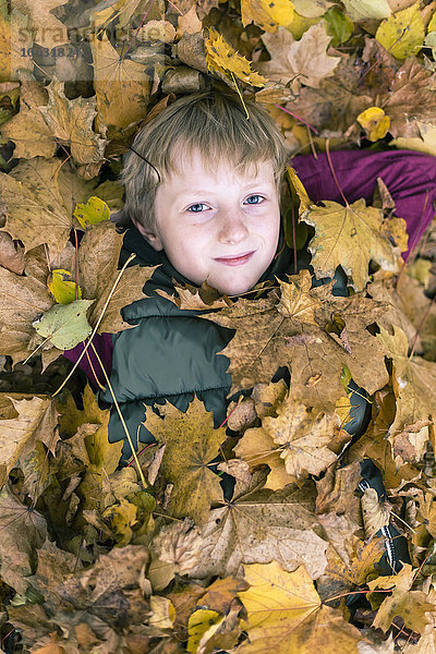 Porträt eines Jungen  der auf dem Boden liegt und mit Herbstblättern bedeckt ist.