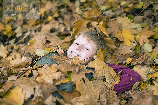 Porträt eines lächelnden Jungen  der auf dem Boden liegt und mit Herbstblättern bedeckt ist.