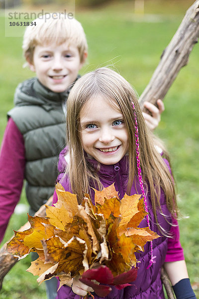 Porträt eines Mädchens mit Herbstblättern  während ihr Bruder im Hintergrund steht.