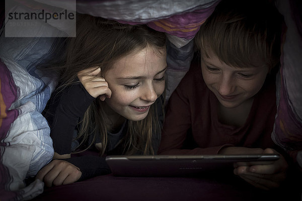Bruder und Schwester liegen nebeneinander und benutzen ein digitales Tablett unter einer Decke.