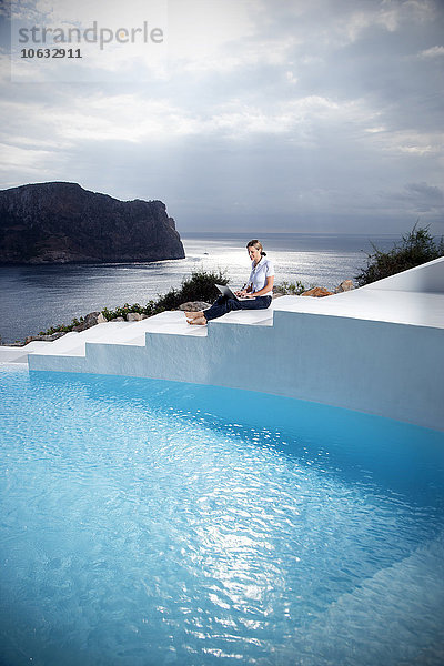 Spanien  Mallorca  Frau mit Laptop auf einer Treppe neben einem Swimmingpool mit Meer im Hintergrund