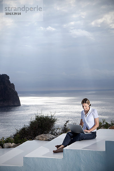 Spanien  Mallorca  Frau mit Laptop auf der Treppe sitzend mit dem Meer im Hintergrund