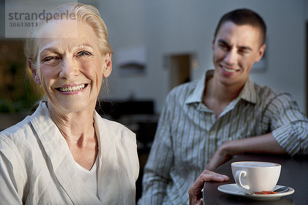 Porträt der lächelnden Seniorin mit Tasse Kaffee und jungem Mann im Hintergrund