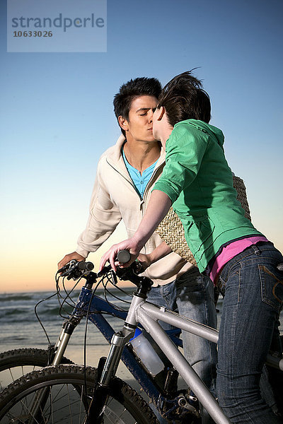 Ein junges Paar  das sich an der Küste auf Fahrrädern küsst.