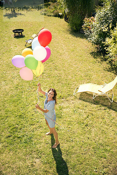 Glückliche Frau steht im Garten und hält bunte Luftballons.