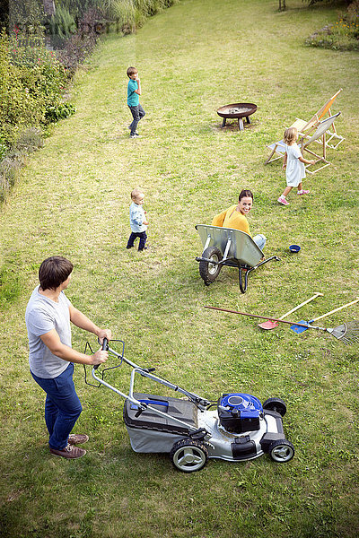 Vater mäht den Rasen im Garten und die Familie spielt im Gras.