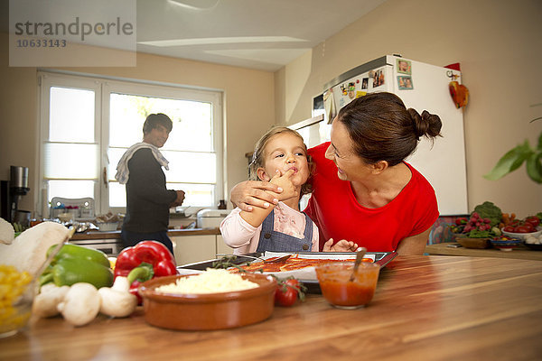 Glückliche Mutter und Tochter in der Küche bereiten Pizza mit Vater im Hintergrund zu