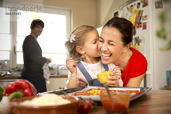 Glückliche Mutter und Tochter in der Küche bereiten Pizza mit Vater im Hintergrund zu