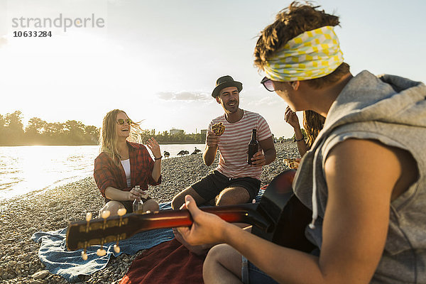 Freunde mit Gitarre entspannen am Flussufer bei Sonnenuntergang
