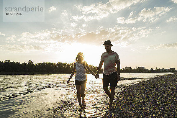 Paar  das bei Sonnenuntergang Hand in Hand am Flussufer spazieren geht.