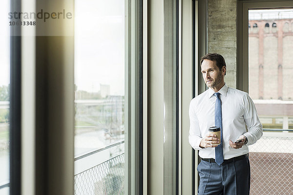 Portrait eines Geschäftsmannes mit Kaffee zum Mitnehmen und Smartphone mit Blick durchs Fenster