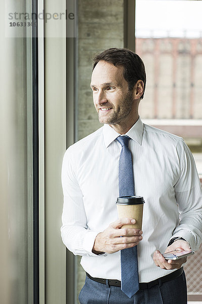 Porträt eines lächelnden Geschäftsmannes mit Kaffee und Smartphone durchs Fenster schauend