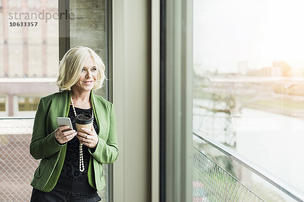 Porträt einer blonden Geschäftsfrau mit Kaffee zum Mitnehmen und Smartphone mit Blick durchs Fenster