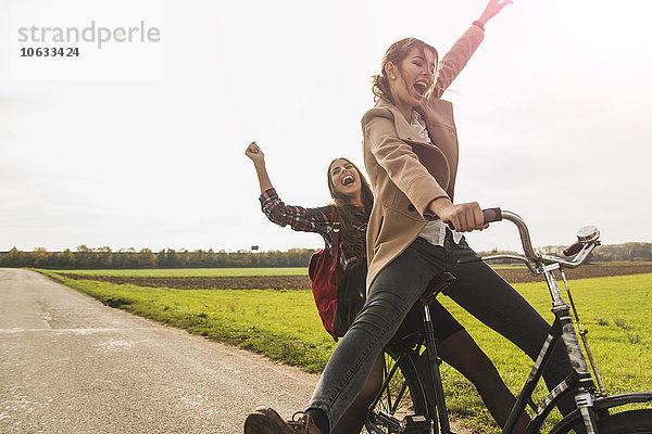 Zwei ausgelassene junge Frauen teilen sich ein Fahrrad in ländlicher Landschaft.