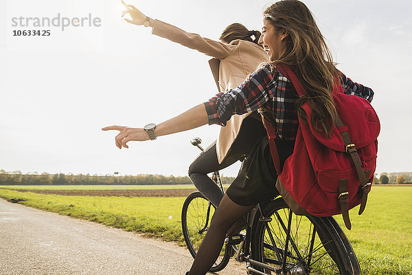 Zwei ausgelassene junge Frauen teilen sich ein Fahrrad in ländlicher Landschaft.