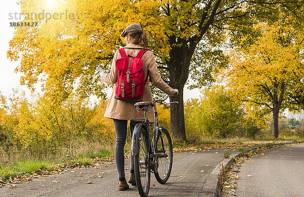 Junge Frau mit Rucksack schiebt ihr Fahrrad in Herbstlandschaft
