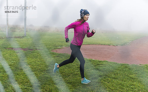 Junge Frau mit Kopfhörer joggen auf einer Wiese hinter Drahtgewebe
