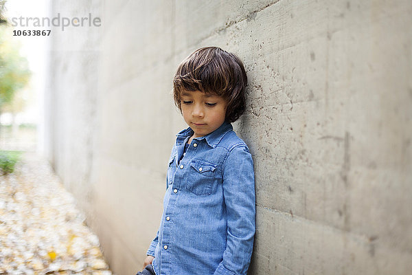 Porträt eines traurigen kleinen Jungen im Jeanshemd  der sich an die Betonwand lehnt.