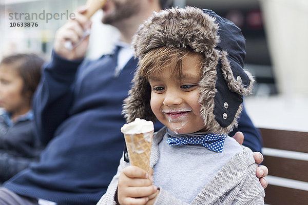 Porträt eines glücklichen kleinen Jungen mit Eistüte  der neben seinem Vater auf einer Bank sitzt.