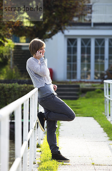 Österreich  Mondsee  lächelnde junge Frau lehnt sich an das Geländer und telefoniert mit dem Smartphone.